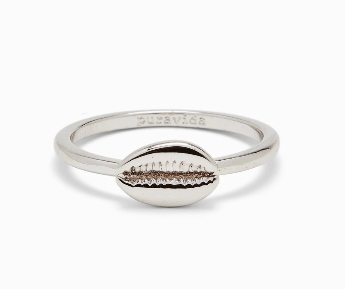 A silver-colored ring with a cowrie shell design on the top, with the phrase 'pura vida' stamped on the inside. Available at Be Charmed