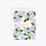 A congratulatory greeting card with a floral pattern and the word 'Congrats' written in a prominent font, accompanied by a white envelope. Available at Be Charmed