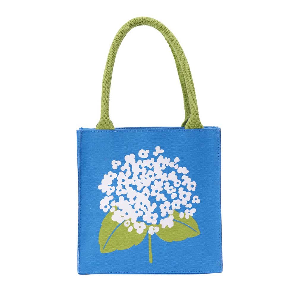 A blue canvas tote bag with light green woven handles and a white flower with green leaves printed on the front, set against a plain white background. Available at Be Charmed