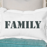 A white rectangular pillow with the word 'Family' printed in black text, placed on a blue couch. Available at Be Charmed