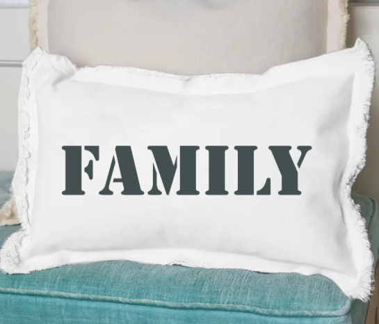 A white rectangular pillow with the word 'Family' printed in black text, placed on a blue couch. Available at Be Charmed