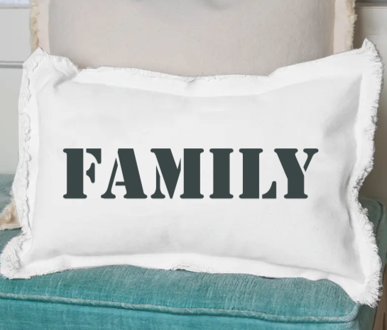 A white rectangular pillow with the word 'Family' printed in black text, placed on a blue couch. Available at Be Charmed
