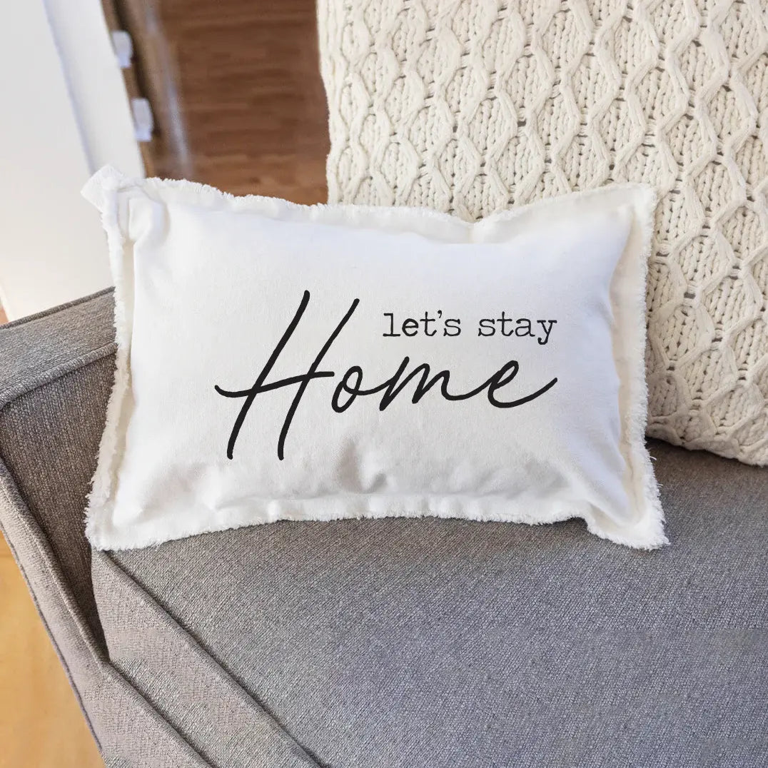 A white rectangular pillow with the text 'Let's Stay Home' printed in black, placed on a couch. Available at Be Charmed