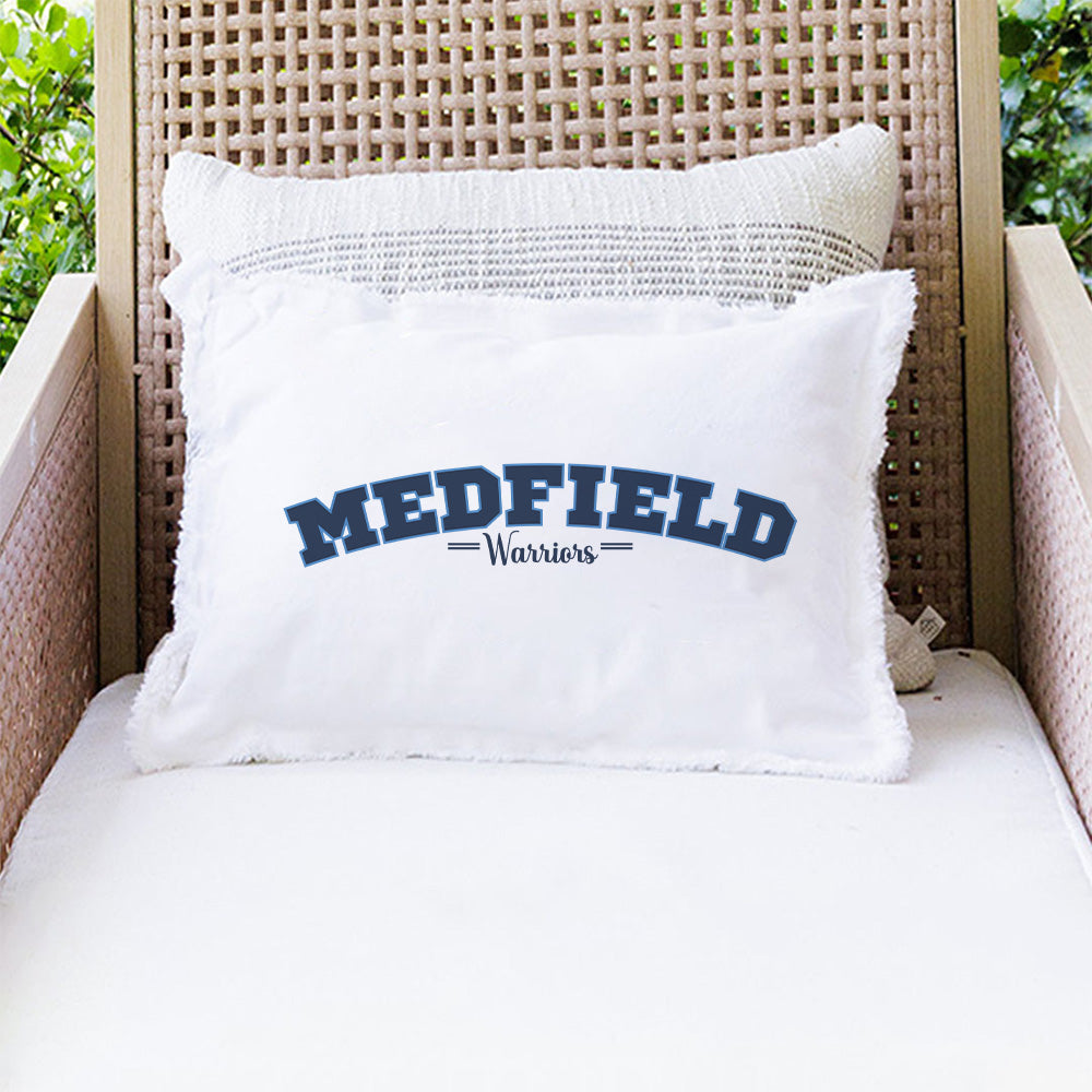 A white lumbar pillow with 'MEDFIELD' and 'Warriors' text in navy blue on a wicker chair. Available at Be Charmed