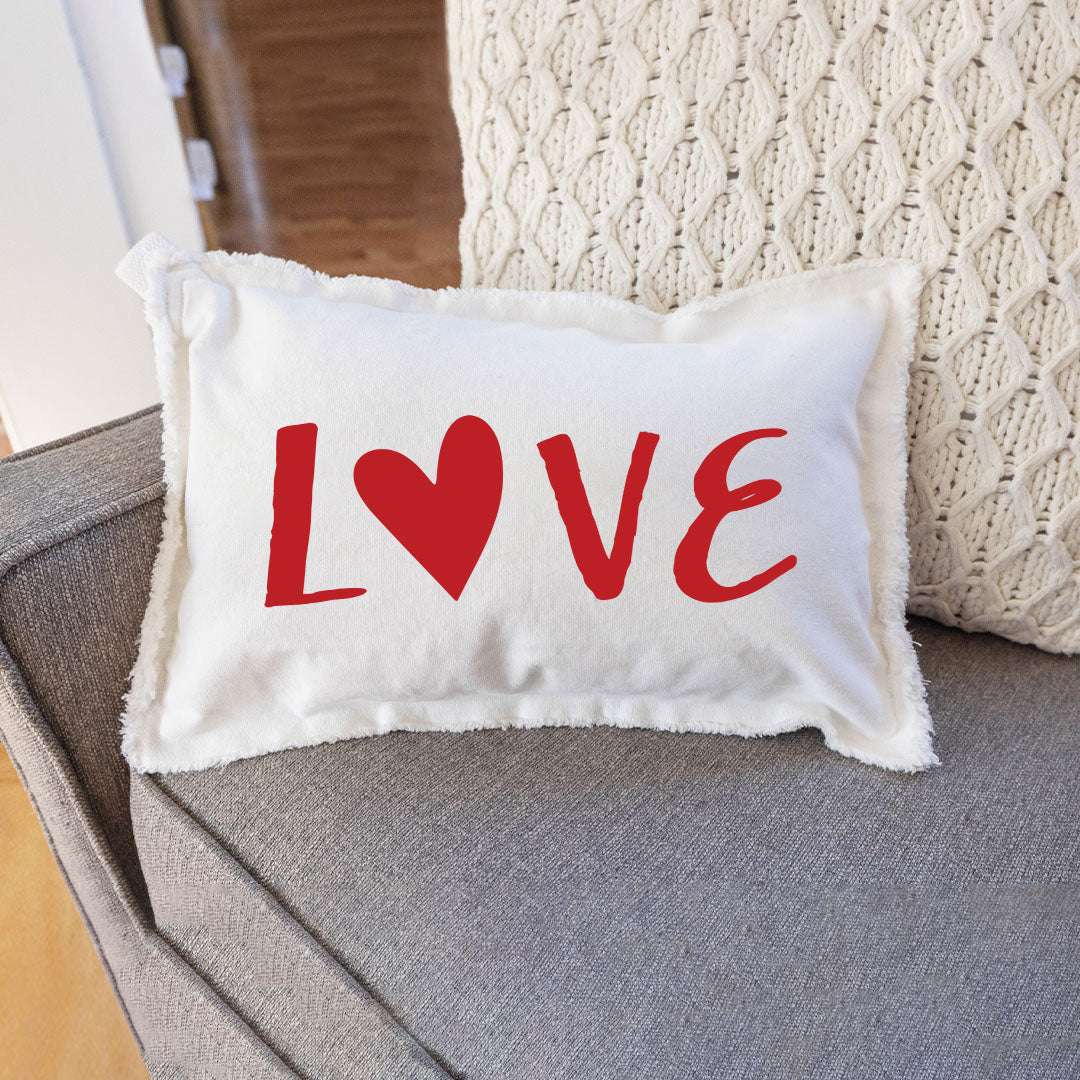 A white rectangular lumbar pillow with a red heart and the word 'love' printed on it, placed on a couch. Available at Be Charmed