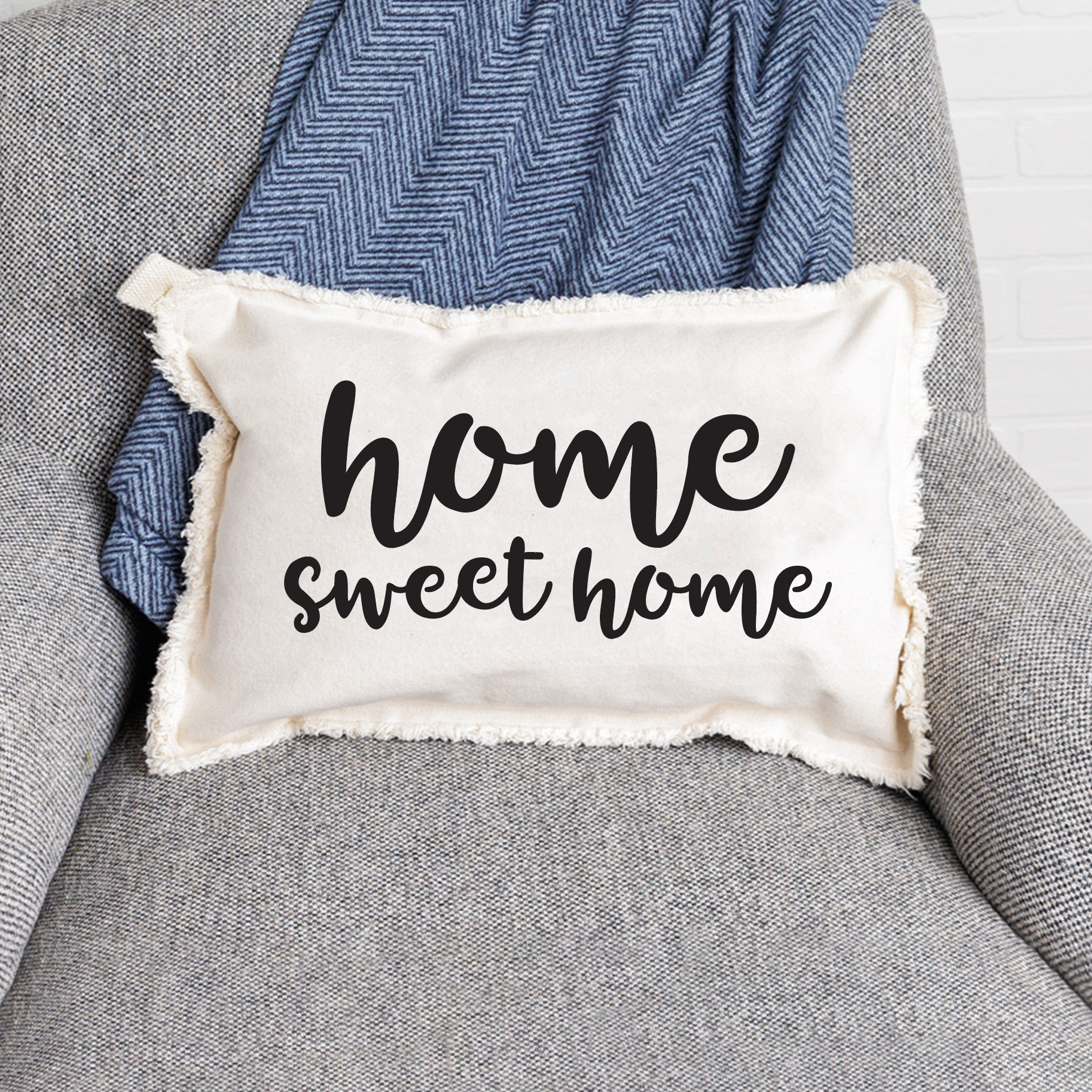 A white rectangular lumbar pillow with the text 'HOME SWEET HOME' written in black script, placed on a gray chair with a blue patterned throw pillow on top. Available at Be Charmed