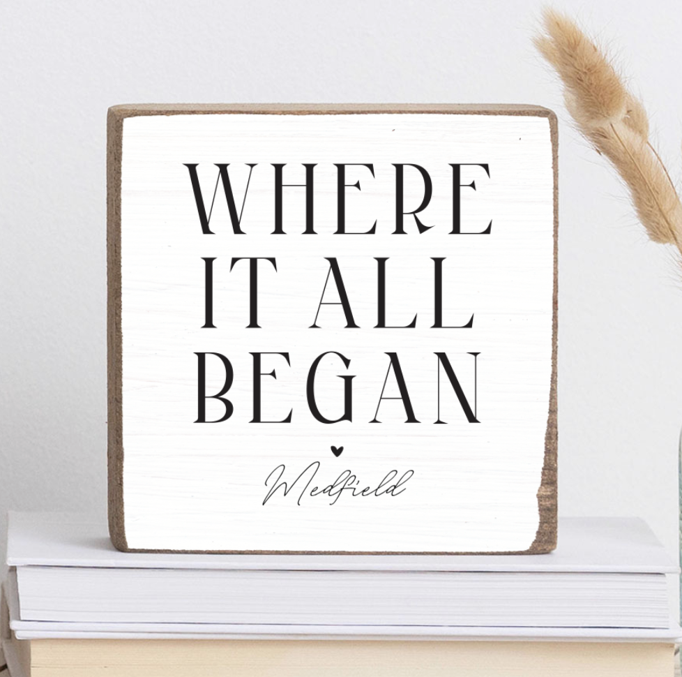 A rustic marlin wooden block with the text 'WHERE IT ALL BEGAN' and 'Medfield' printed on it, displayed on a stack of books. Availble at Be Charmed
