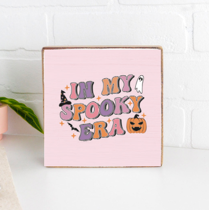 A decorative wooden block with a pink background, featuring the text 'In My Spooky Era' accompanied by an illustration of a white ghost, a pumpkin, and some candy. Available at Be Charmed