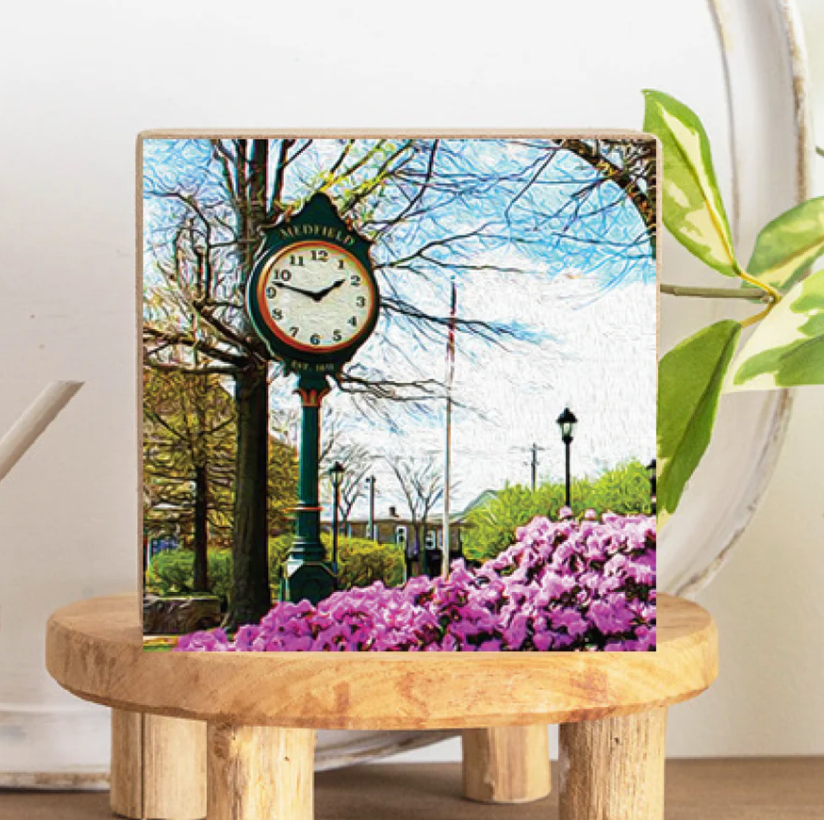 A decorative wooden block featuring an image of a clock on a pedestal surrounded by trees and flowers, displayed on a wooden stool. Available at Be Charmed