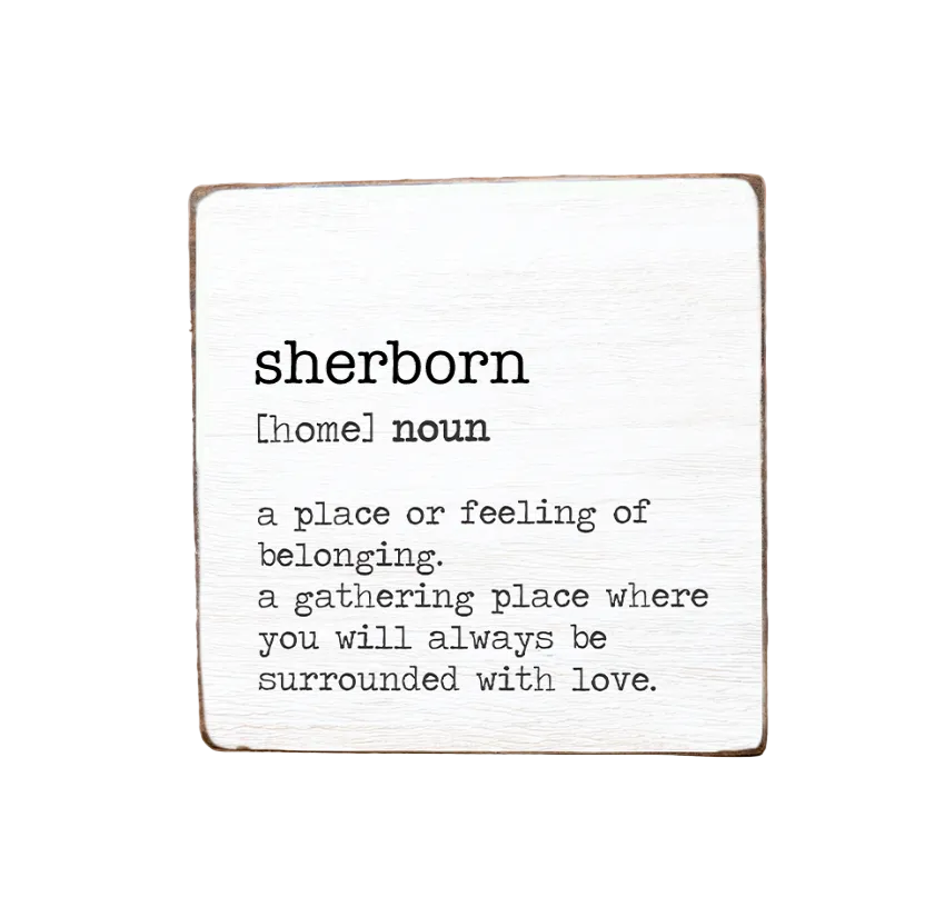 A wooden block with the text 'SHERBORN' and a definition of the word 'HOMELAND' written on it, placed on a wooden surface with a floral pattern in the background. Available at Be Charmed