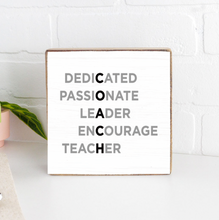 A wooden block sign with the word 'DEDICATED' at the top, and the words 'Passionate', 'Leader', 'Encourage', 'Teacher' below it, placed on a white surface with a brick wall in the background and a potted plant to the side. Available at Be Charmed