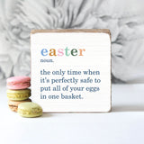 A decorative wooden block with the text 'Easter' at the top, and the phrase 'The only time when it’s perfectly safe to put all of your eggs in one basket.' below it, accompanied by a set of multi-colored macaroon cookies to the side. Available at Be Charmed