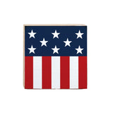 A decorative wooden block sign featuring the American flag in red, white, and blue colors, placed on a surface with a blue patterned background. Available at Be Charmed