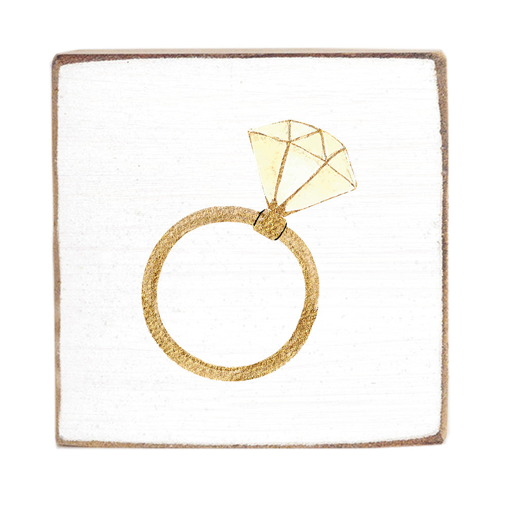 A gold-colored ring-shaped solitaire brooch displayed on a white square rustic block. Available at Be Charmed