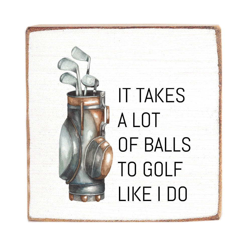 A 6x6 wooden block with a rustic design featuring an illustration of golf clubs and a golf bag, and the text 'IT TAKES A LOT OF BALLS TO GOLF LIKE I DO'. Available at Be Charmed
