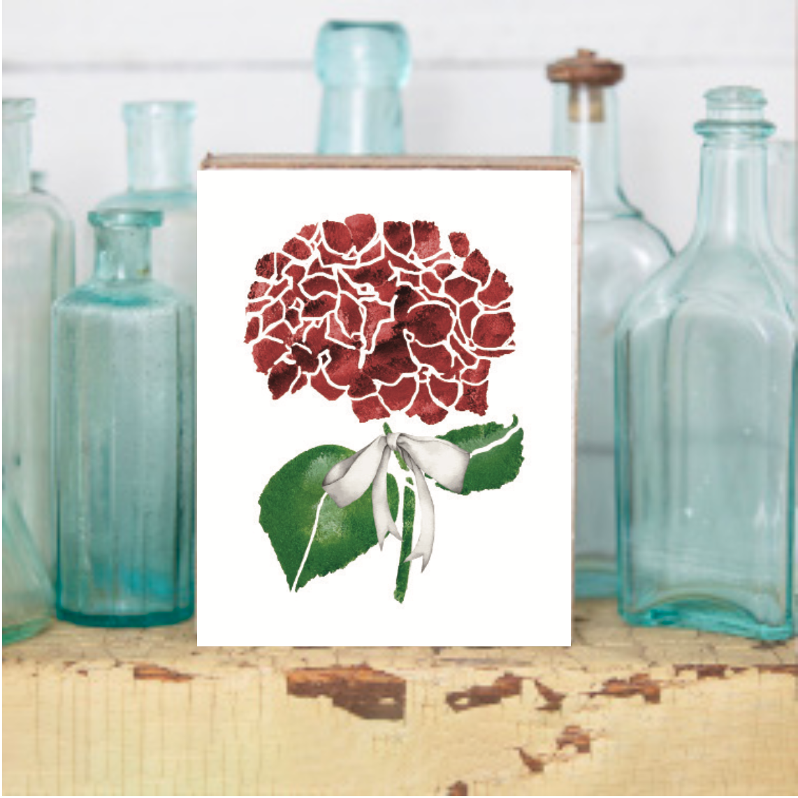 A decorative wooden block featuring a print of a red and green hydrangea flower with a white ribbon, placed on a wooden surface with glass bottles in the background. Available at Be Charmed