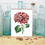 A decorative wooden block featuring a print of a red and green hydrangea flower with a white ribbon, placed on a wooden surface with glass bottles in the background. Available at Be Charmed