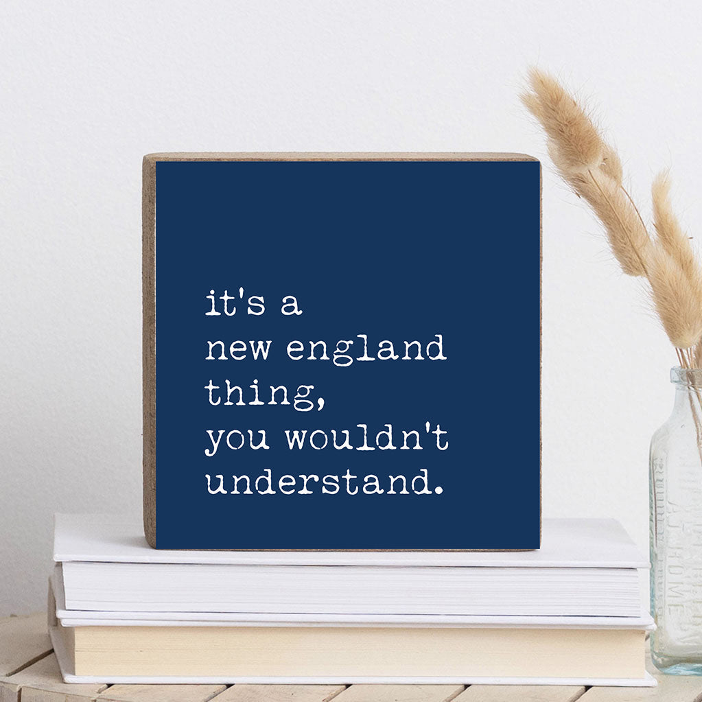 A navy blue Rustic Marlin wooden block with white text 'it's a new england thing, you wouldn't understand' on it, displayed on a stack of books. Available at Be Charmed
