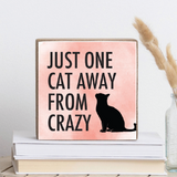 A wooden decorative block with a cat silhouette and the phrase 'JUST ONE CAT AWAY FROM CRAZY' on a pink background, placed on a stack of books. Available at Be Charmed