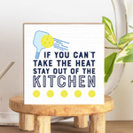 A decorative wooden block with a pickleball paddle design and the text 'IF YOU CAN'T TAKE THE HEAT STAY OUT OF THE KITCHEN' on a white background, placed on a wooden stand. Available at Be Charmed