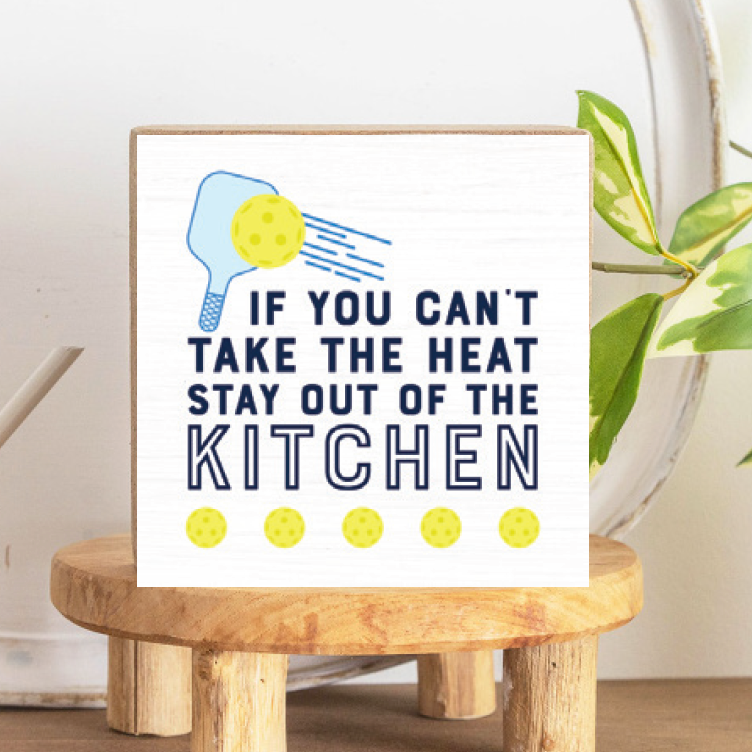 A decorative wooden block with a pickleball paddle design and the text 'IF YOU CAN'T TAKE THE HEAT STAY OUT OF THE KITCHEN' on a white background, placed on a wooden stand. Available at Be Charmed