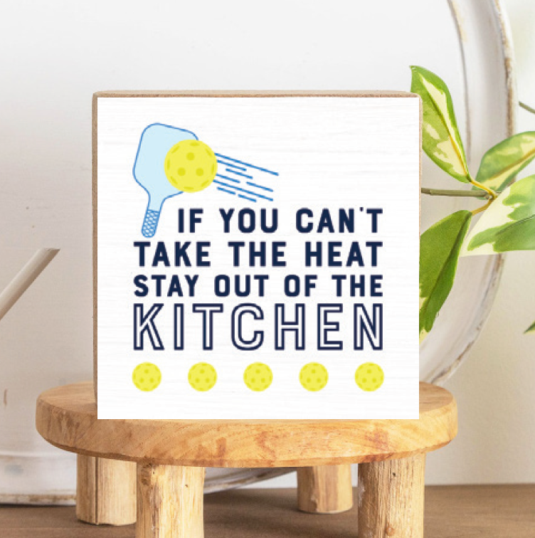 A decorative wooden block with a pickleball paddle design and the text 'IF YOU CAN'T TAKE THE HEAT STAY OUT OF THE KITCHEN' on a white background, placed on a wooden stand. Available at Be Charmed