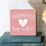 A rectangular wooden block with pink background, text that reads 'Promoted to Big Sister' and a heart symbol, placed on a surface with a vase of pink flowers in the background. Available at Be Charmed