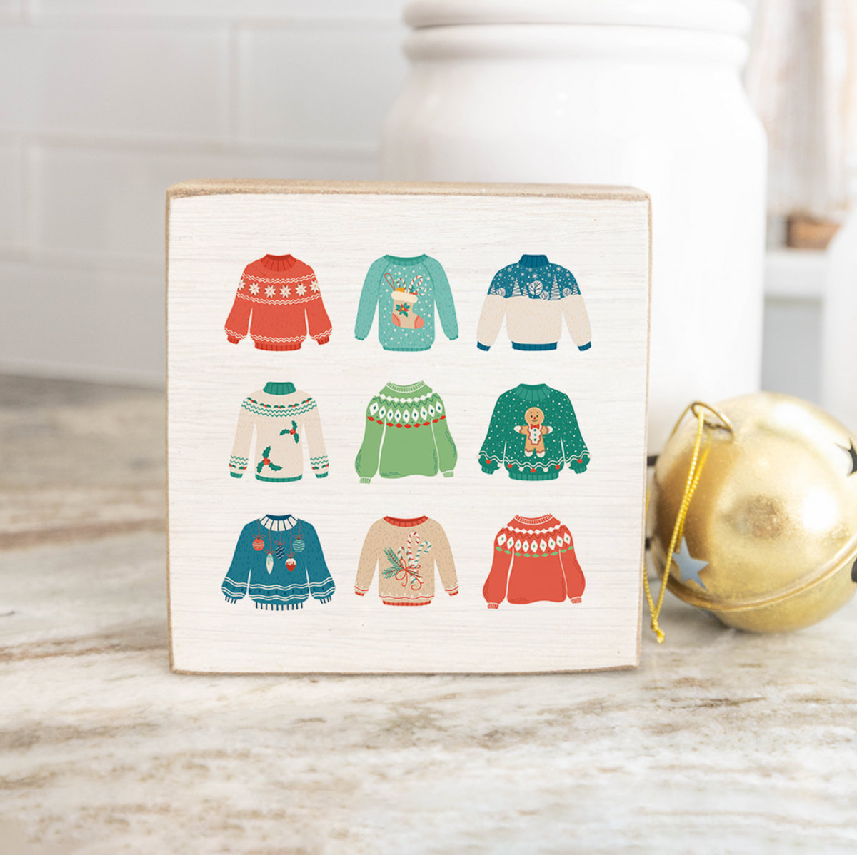 A decorative wooden block with various colorful Christmas sweater designs illustrated on it, placed on a marble surface next to a gold-colored ornament and a white vase with a Christmas tree. Available at Be Charmed