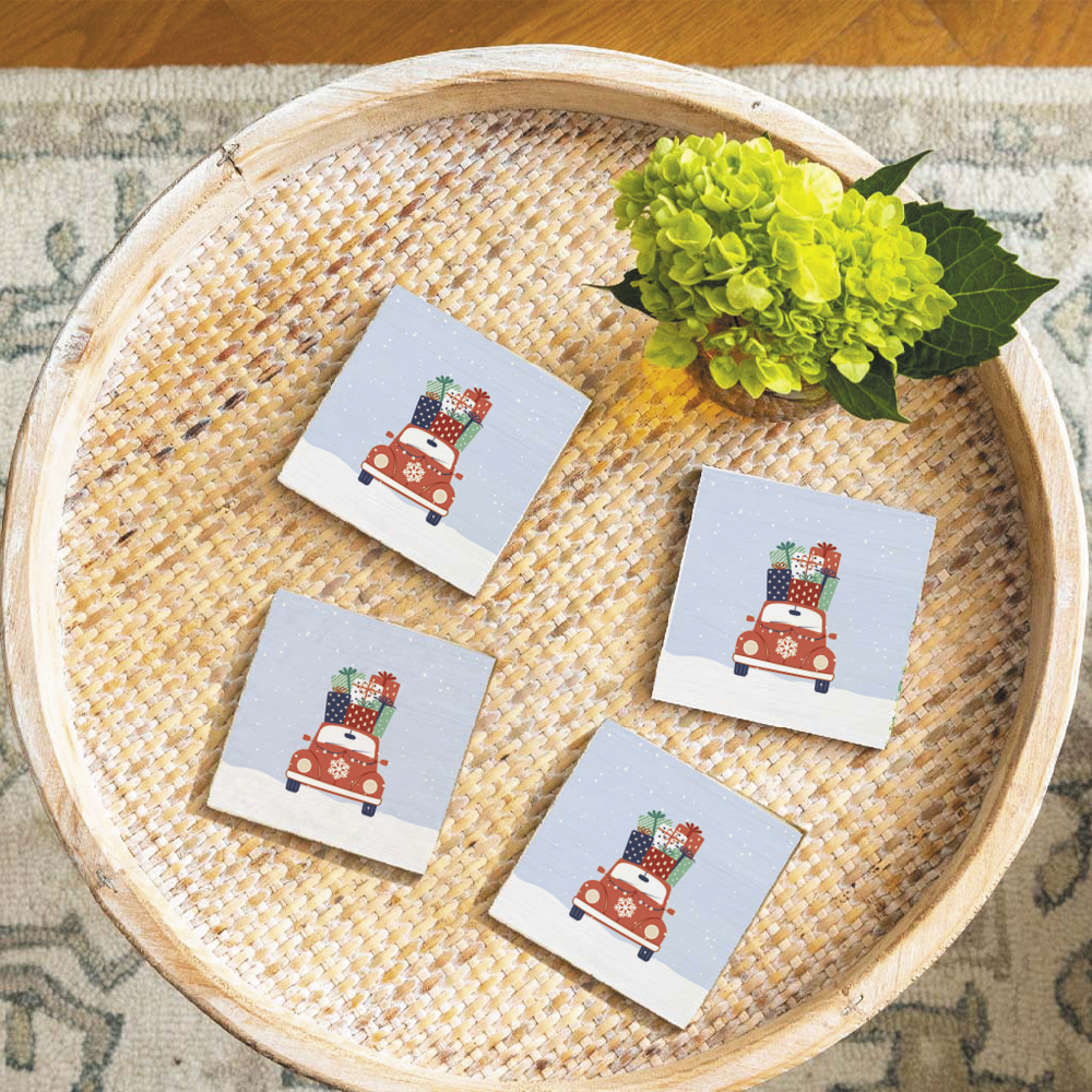 A set of four round solid wood coasters with a rustic design featuring a red car loaded with gifts, placed on a woven mat. Available at Be Charmed