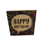 A decorative Rustic Marlin wooden block with 'HAPPY BIRTHDAY!' written in black on a beige background, surrounded by a multicolored confetti pattern. Available at Be Charmed