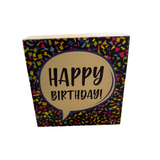 A decorative Rustic Marlin wooden block with 'HAPPY BIRTHDAY!' written in black on a beige background, surrounded by a multicolored confetti pattern. Available at Be Charmed