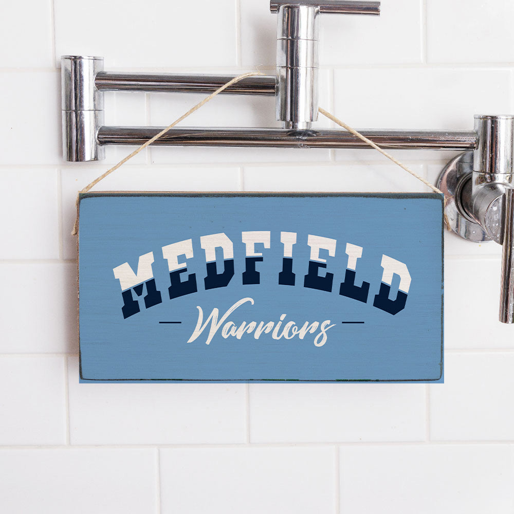 A rectangular blue twine sign with the text 'MEDFIELD Warriors' hanging against a white tiled background with silver pipes. Available at Be Charmed