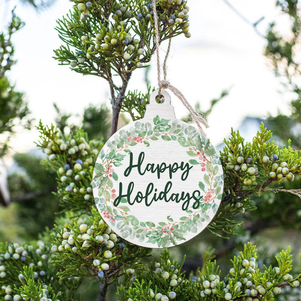 A handcrafted wooden ornament with a 'Happy Holidays' message, hanging from a tree with green foliage in the background. Available at Be Charmed