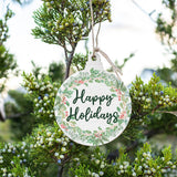 A handcrafted wooden ornament with a 'Happy Holidays' message, hanging from a tree with green foliage in the background. Available at Be Charmed