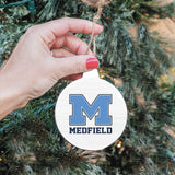 A hand holding a round, white Christmas ornament with the letter 'M' in the center, against a backdrop of green Christmas trees. Available at Be Charmed