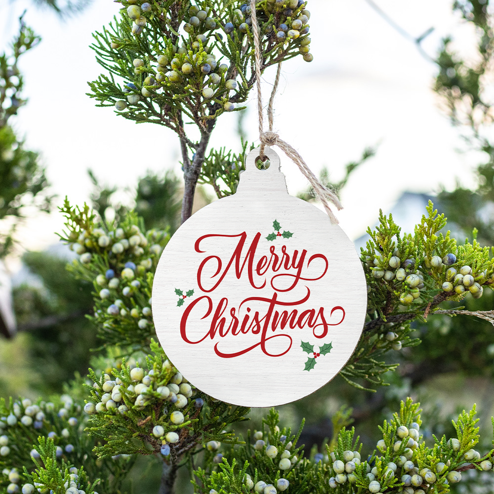 A rustic wooden bulb ornament with the text 'Merry Christmas' and holly leaves, hanging from a branch with a natural background. Available at Be Charmed