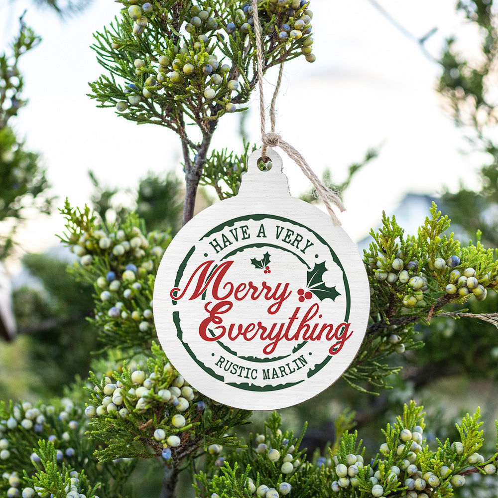 A round, handcrafted wooden ornament with a 'Merry Everything' text and a rustic marlin design, hanging from a branch with greenery in the background. Available at Be Charmed