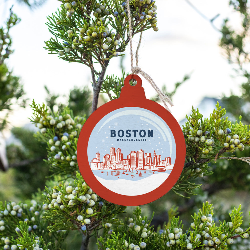 A round, red, white, and blue ornament with 'Boston, Massachusetts' written on it, hanging from a branch with green foliage in the background. Available at Be Charmed