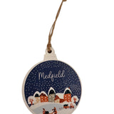 A round, rustic wooden ornament with a bulb design, featuring a vintage snow globe village scene in blue and white colors, with the word 'MEDField' written on it. Available at Be Charmed
