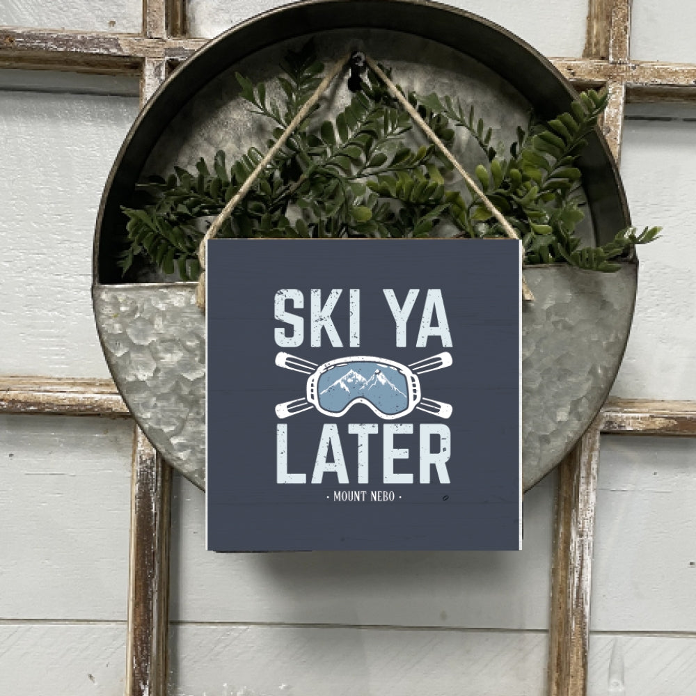 A square wooden sign with the phrase 'SKI YA LATER' in bold letters, surrounded by an illustration of skis and mountains, hung by twine. Available at Be Charmed