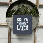 A square wooden sign with the phrase 'SKI YA LATER' in bold letters, surrounded by an illustration of skis and mountains, hung by twine. Available at Be Charmed