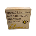 A beige wooden block with the phrase 'spread kindness like a honeybee and watch the world bloom' printed on it, along with an image of a honeybee. Available at Be Charmed