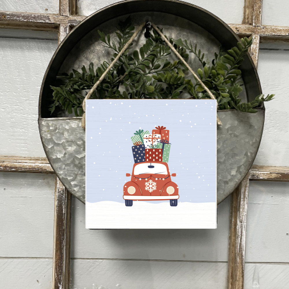 A square wooden twine sign with a holiday haul theme, featuring a red car with gifts on a snowy background, hung on a wall with a round mirror and green foliage in the background. Available at Be Charmed