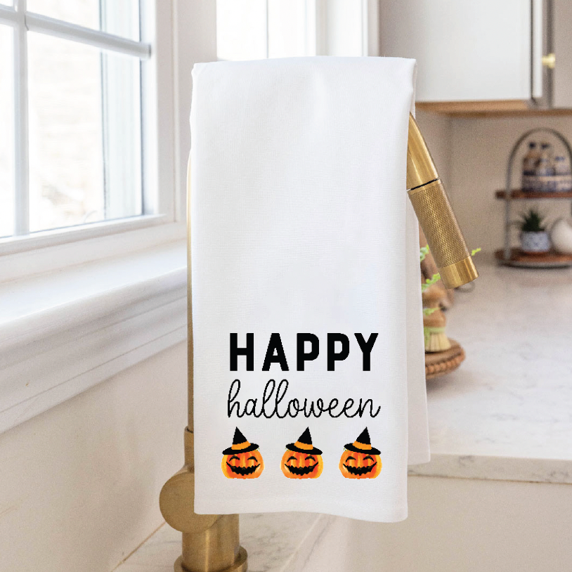 A white tea towel with 'Happy Halloween' text and decorative pumpkin designs hanging on a bronze kitchen towel holder. Available at Be Charmed