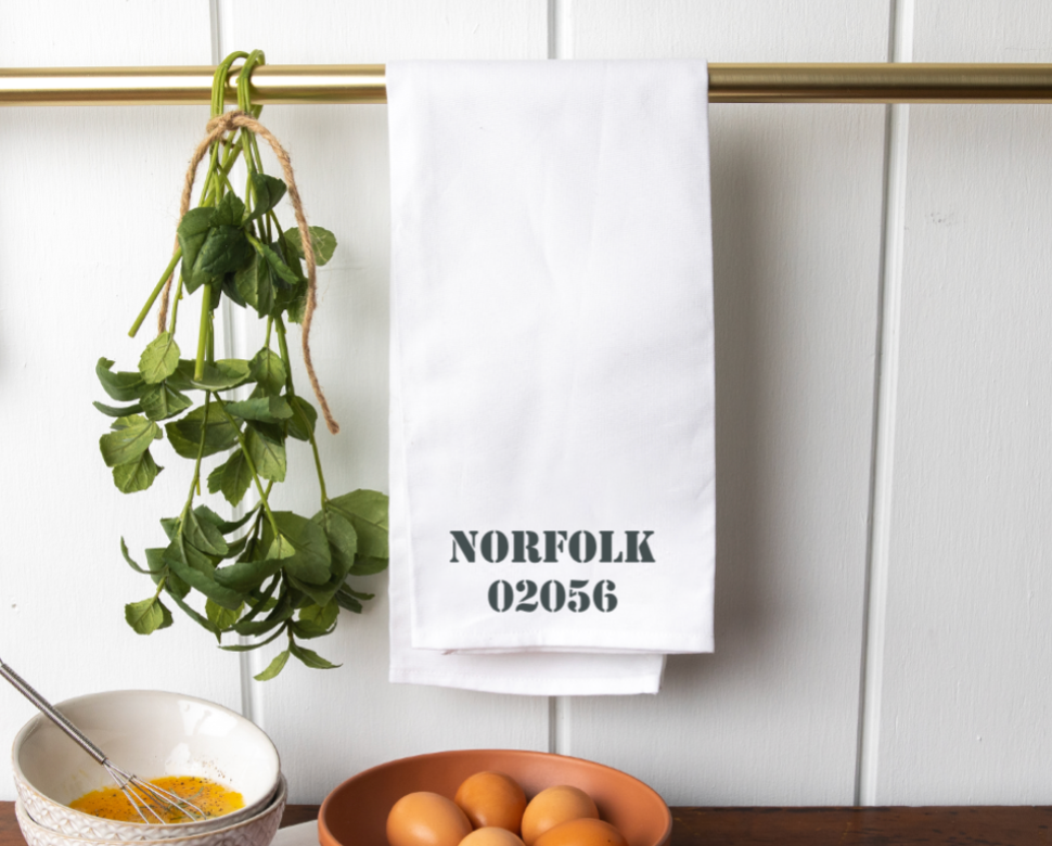 A white tea towel with the text 'NORFOLK 02056' and a small green plant, hanging on a wall hook. Availablat at Be Charmed