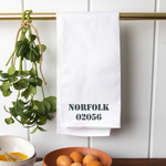A white tea towel with the text 'NORFOLK 02056' and a small green plant, hanging on a wall hook. Availablat at Be Charmed