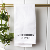 A white tea towel with the name 'Sherborn' and a number '0177' printed on it, hanging on a gold-colored rail with green foliage in the background. Available at Be Charmed