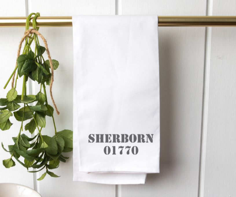 A white tea towel with the name 'Sherborn' and a number '0177' printed on it, hanging on a gold-colored rail with green foliage in the background. Available at Be Charmed