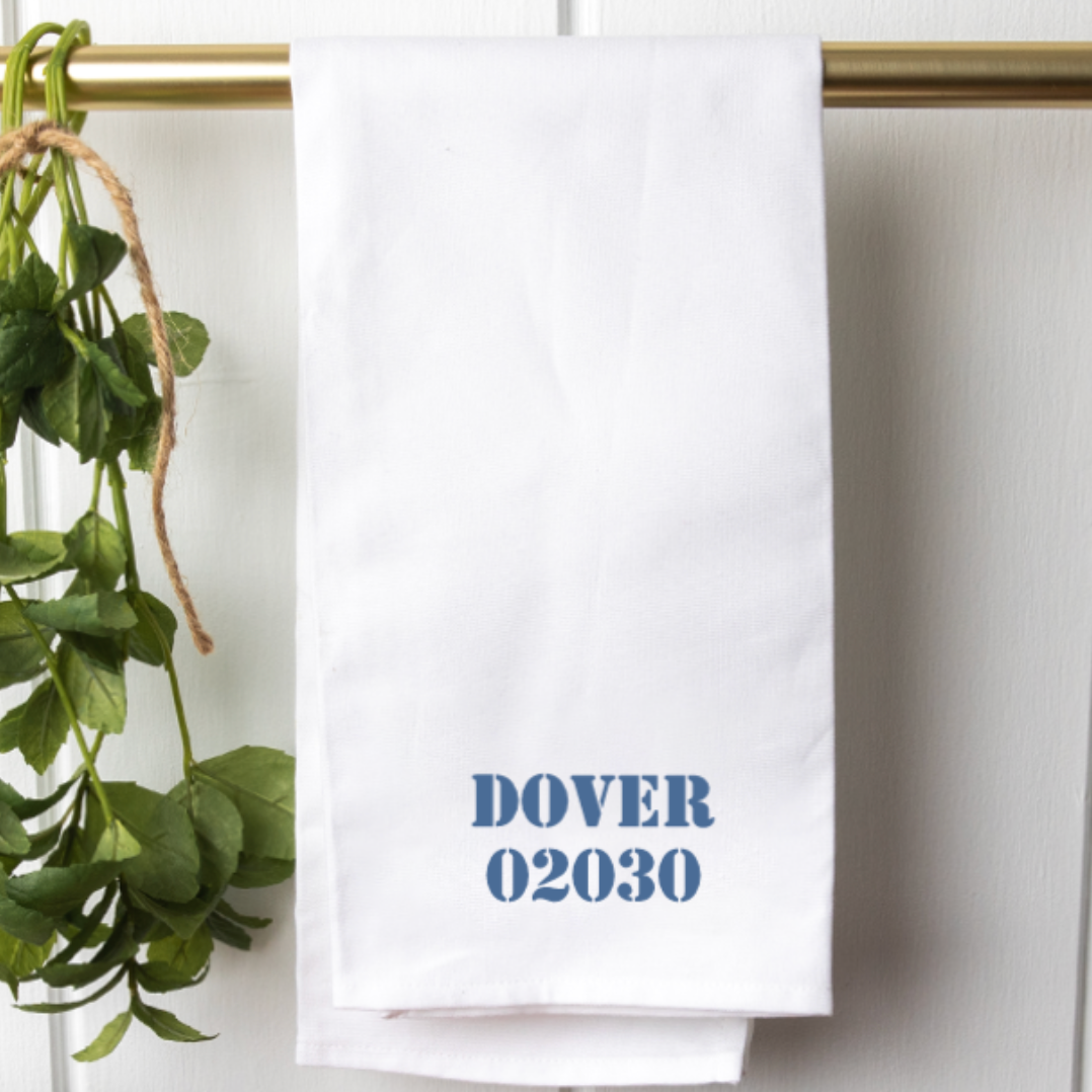 A white cotton tea towel with the text 'DOVER 02030' printed in blue ink, displayed on a banister alongside a green plant. Available at Be Charmed