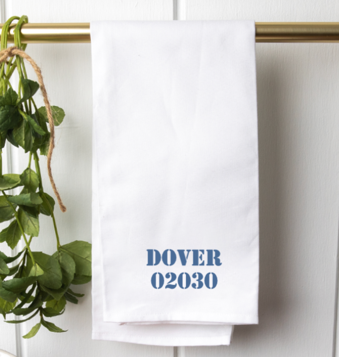 A white cotton tea towel with the text 'DOVER 02030' printed in blue ink, displayed on a banister alongside a green plant. Available at Be Charmed